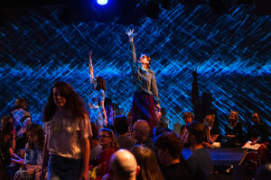 A group of people are on stage during an event; some participants raise their hands, with blue light projections in the background.